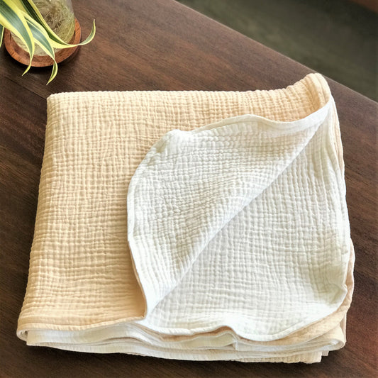 a folded beige blanket or quilt placed on a wooden surface, with a small potted plant visible in the background.