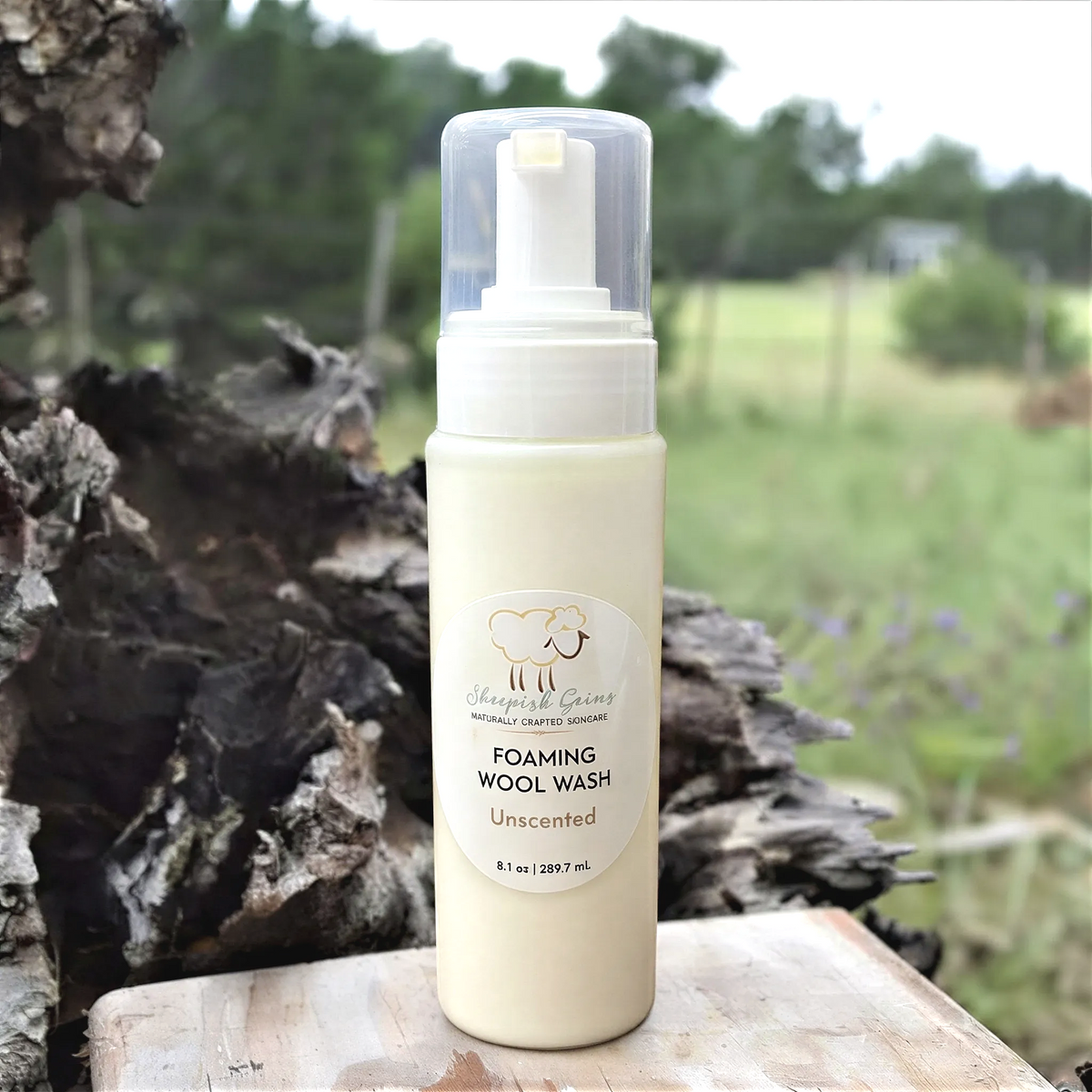 A bottle of Sheepish Gains Foaming Wool Wash Unscented, with a label that reads "Unscented" and "8 fl oz (1.39 fl oz)". The bottle is placed on a wooden surface, with a blurred background of trees and grass.